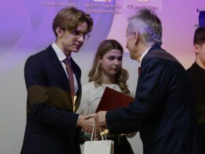 Zdjęcie pokazuje moment wręczenia nagrody. Młody mężczyzna w garniturze, stojący po lewej stronie, ściska dłoń starszego mężczyzny w garniturze po prawej. Młodemu mężczyźnie towarzyszy młoda kobieta stojąca za nim, trzymająca czerwoną teczkę. W ręku młodego mężczyzny widać jasną torebkę upominkową. Tło stanowi ekran z lekkim fioletowym podświetleniem.