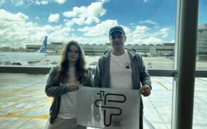 Na zdjęciu stoją obok siebie młoda kobieta i młody mężczyzna. Oboje patrzą prosto w obiektyw i trzymają rozłożoną flagę szkolną z logotypem. Oboje mają na sobie jasne bluzy w podobnym odcieniu szarości. Mężczyzna nosi ciemną czapkę z daszkiem. Za nimi, przez duże panoramiczne okno, widać płytę lotniska, biały samolot pasażerski oraz terminale lotnicze. Nad wszystkim rozciąga się jasne niebo z licznymi białymi chmurami. Zdjęcie sprawia wrażenie dokumentującego początek wyprawy lub powrót z zawodów.