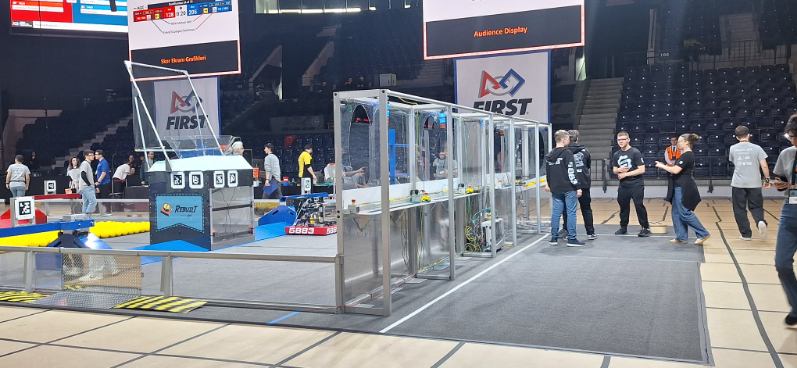 Wnętrze dużej hali sportowej podczas zawodów robotycznych FIRST. Na środku znajduje się prostokątne pole gry otoczone niską metalową barierką i przezroczystymi panelami ochronnymi. Na planszy leżą liczne żółte piłki. Wokół pola stoją uczestnicy i organizatorzy w koszulkach drużynowych. Nad areną wiszą duże ekrany z wynikami i logotypem FIRST.