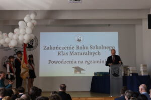Zdjęcie przedstawia uroczystość zakończenia roku szkolnego klas maturalnych odbywającą się w sali szkolnej. Na środku sceny wyświetlany jest duży biały ekran z napisem: „Zakończenie Roku Szkolnego Klas Maturalnych. Powodzenia na egzaminie”. Pod napisem znajduje się grafika czapki absolwenta. Po prawej stronie stoi mężczyzna w garniturze przy mównicy, prawdopodobnie przemawiający dyrektor lub nauczyciel. Obok ustawiono stosy książek lub nagród oraz rząd kolorowych upominków. Po lewej stronie znajdują się uczniowie pocztu sztandarowego w galowych strojach – białe koszule, ciemne spódnice lub spodnie oraz biało-czerwone elementy. Jeden z uczniów trzyma sztandar szkoły. Nad nimi widoczna jest dekoracja z białych balonów. Na dole zdjęcia widać siedzącą publiczność obserwującą uroczystość. Całość ma oficjalny i podniosły charakter związany z zakończeniem nauki przez maturzystów.