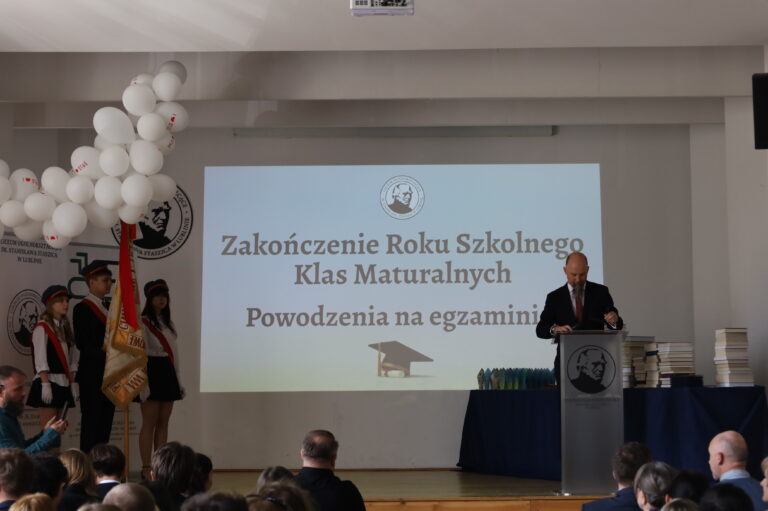 Zdjęcie przedstawia uroczystość zakończenia roku szkolnego klas maturalnych odbywającą się w sali szkolnej. Na środku sceny wyświetlany jest duży biały ekran z napisem: „Zakończenie Roku Szkolnego Klas Maturalnych. Powodzenia na egzaminie”. Pod napisem znajduje się grafika czapki absolwenta. Po prawej stronie stoi mężczyzna w garniturze przy mównicy, prawdopodobnie przemawiający dyrektor lub nauczyciel. Obok ustawiono stosy książek lub nagród oraz rząd kolorowych upominków. Po lewej stronie znajdują się uczniowie pocztu sztandarowego w galowych strojach – białe koszule, ciemne spódnice lub spodnie oraz biało-czerwone elementy. Jeden z uczniów trzyma sztandar szkoły. Nad nimi widoczna jest dekoracja z białych balonów. Na dole zdjęcia widać siedzącą publiczność obserwującą uroczystość. Całość ma oficjalny i podniosły charakter związany z zakończeniem nauki przez maturzystów.